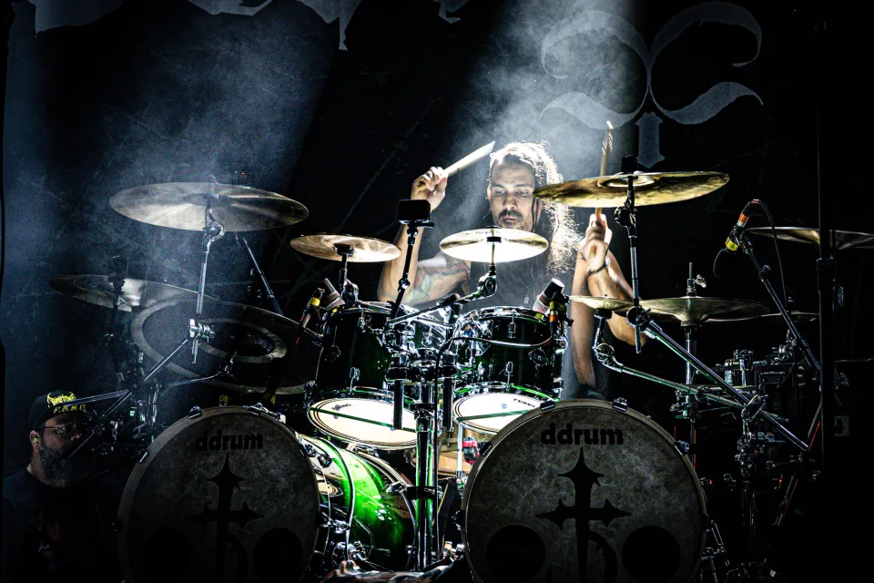 Drummer surrounded by cymbals, backlit by a beam of light through stage smoke.